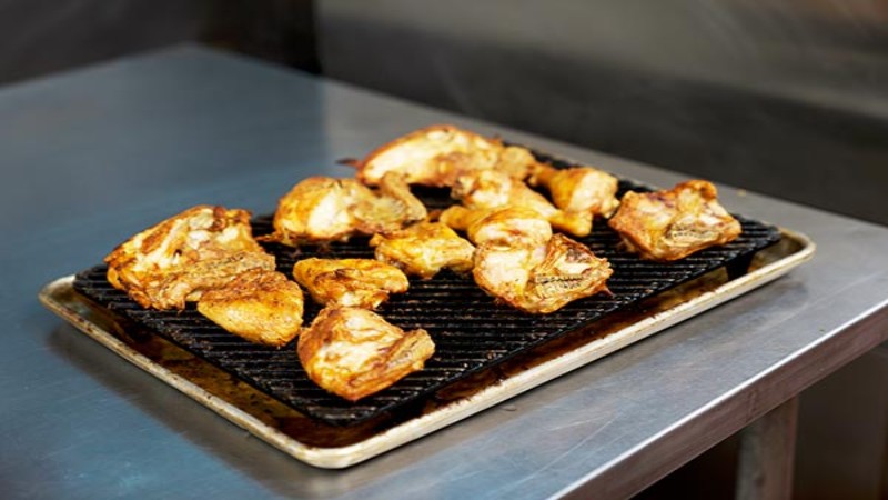 Grilled chicken pieces on rack placed on baking tray in commercial kitchen.