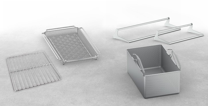 An assortment of stainless steel cooking accessories including a perforated tray, a flat rack, and a container, displayed on a light surface.