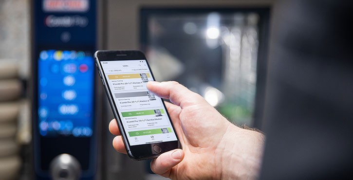 A hand holding a smartphone in front of an iCombi Pro with visible display in the kitchen.