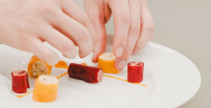 Gros plan sur les mains d’un chef disposant soigneusement des rouleaux de légumes colorés sur une assiette blanche.