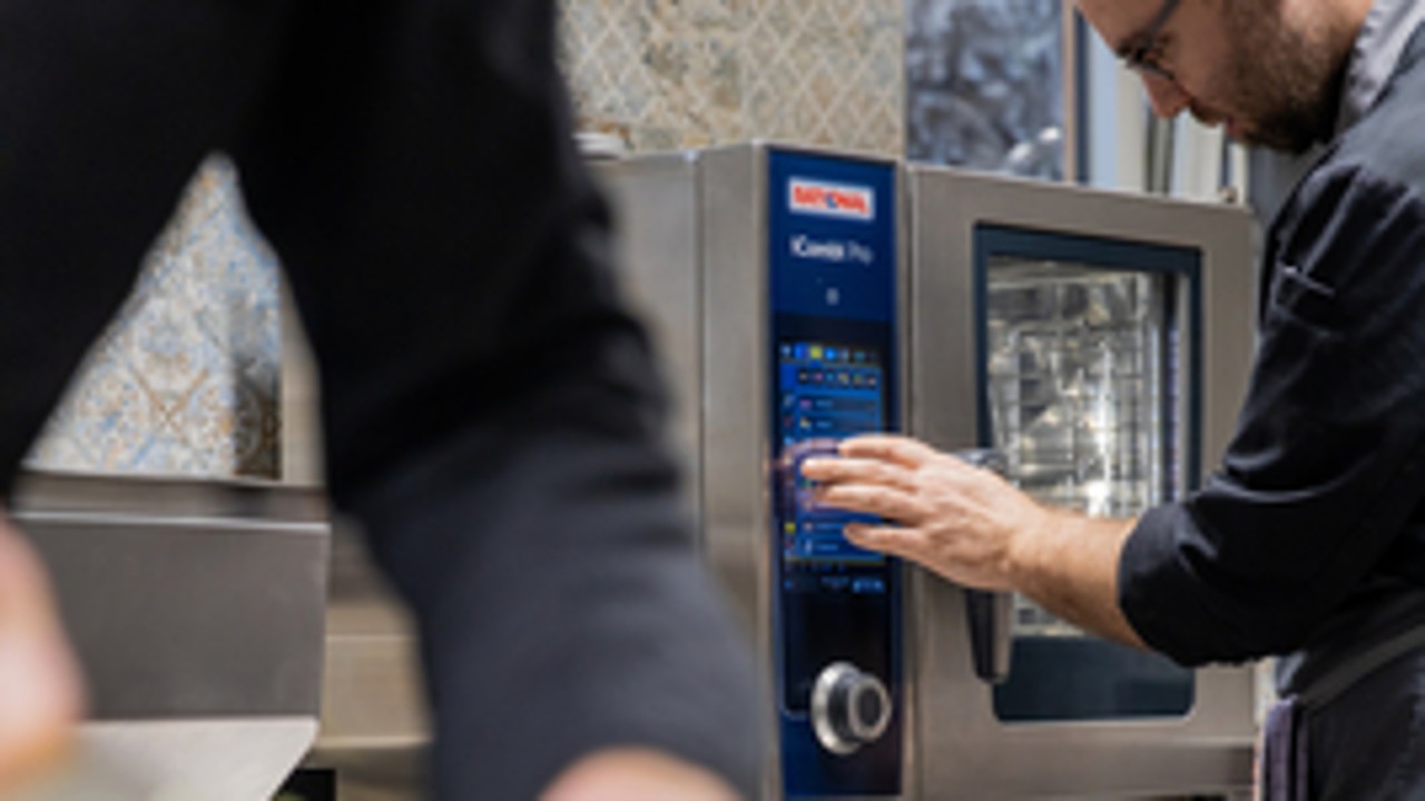 Chef operating a RATIONAL iCombi Pro XS combi oven while a colleague prepares vegetables.