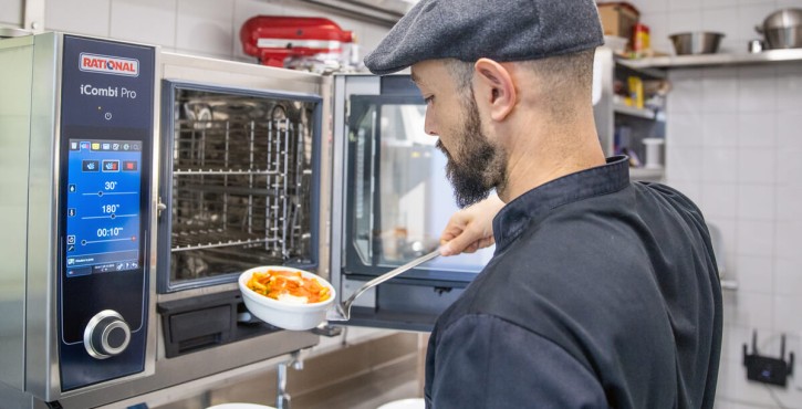 A chef using a Rational iCombi Pro appliance to serve food from a bowl onto a plate in a professional kitchen.