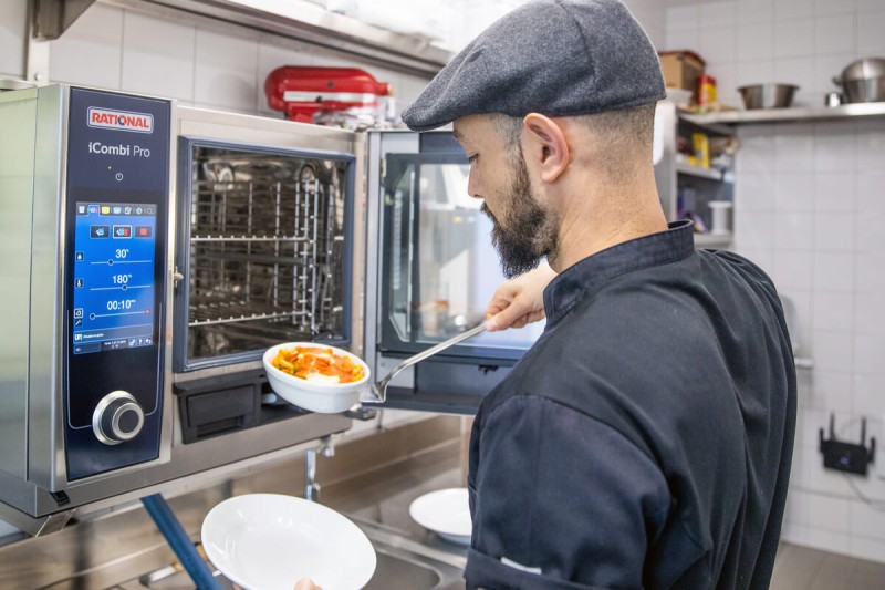 A chef using a Rational iCombi Pro appliance to serve food from a bowl onto a plate in a professional kitchen.