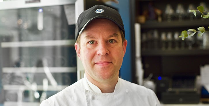 Chef cuisinier en veste blanche et casquette noire dans une cuisine professionnelle.
