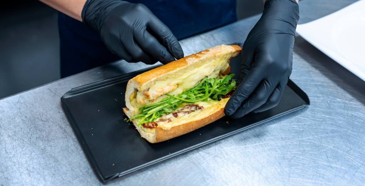 Mani con guanti preparano panino gourmet con formaggio fuso, rucola e pomodori secchi.
