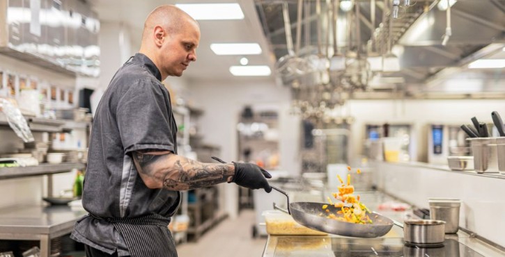 Chef sautéing vegetables in a pan on induction hob in a professional kitchen.