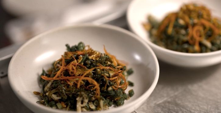 Bowl of sautéed leafy greens with shredded carrots and herbs on stainless steel counter.