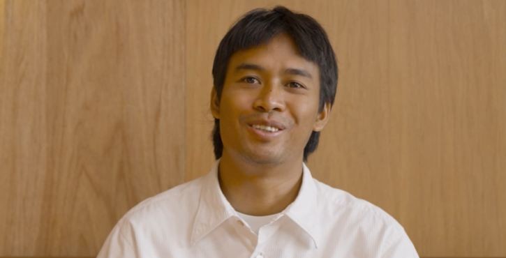 Smiling man in white shirt sitting against wooden wall background.