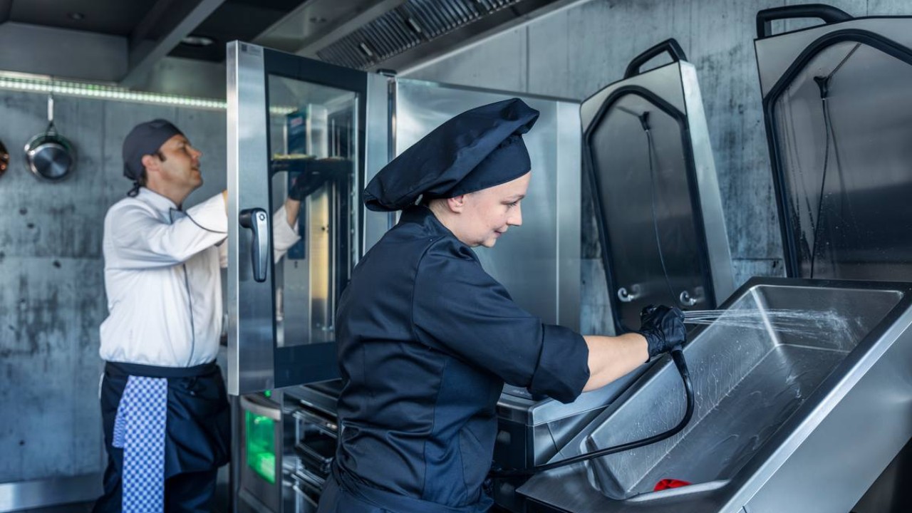 Kuchařka oplachuje RATIONAL iVario Pro hadicí, zatímco další kuchař pracuje s RATIONAL iCombi Pro v moderní kuchyni.