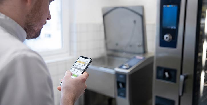 Ein Koch in weißer Uniform, der auf ein Smartphone schaut, während er neben Kochgeräten in einer Küche steht.
