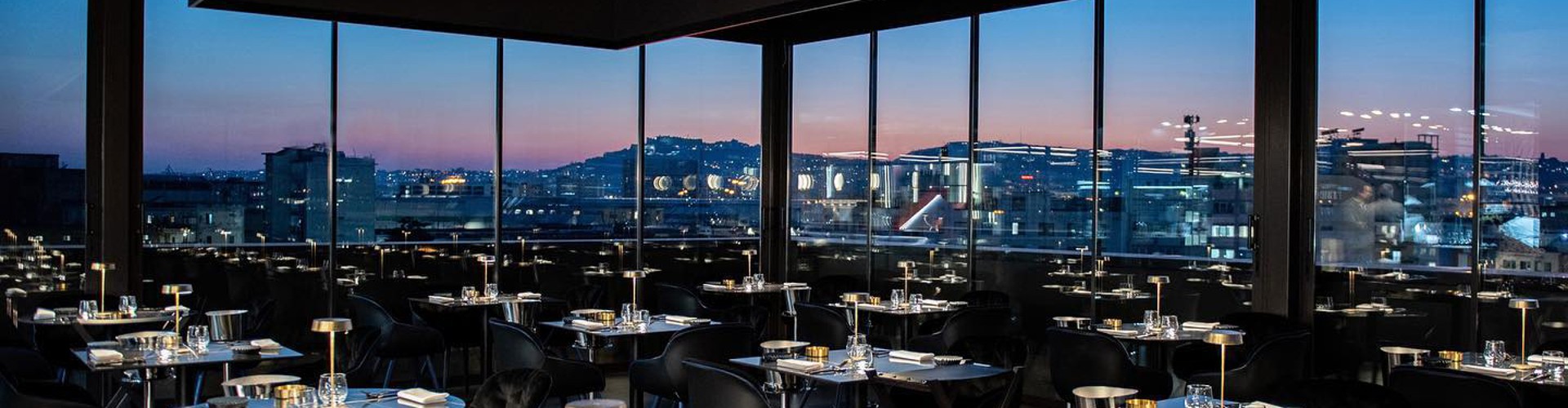 Ristorante panoramico rooftop con vista sulla città attraverso ampie vetrate al tramonto.