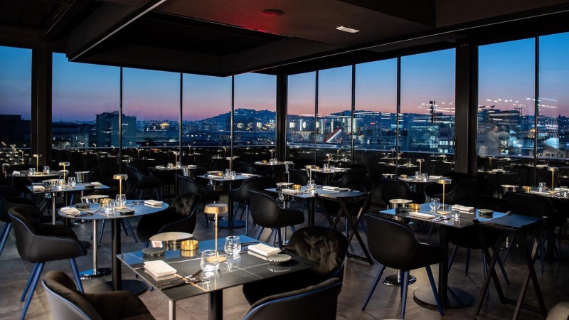 Elegante ristorante rooftop con vista panoramica sulla città attraverso ampie vetrate al tramonto.