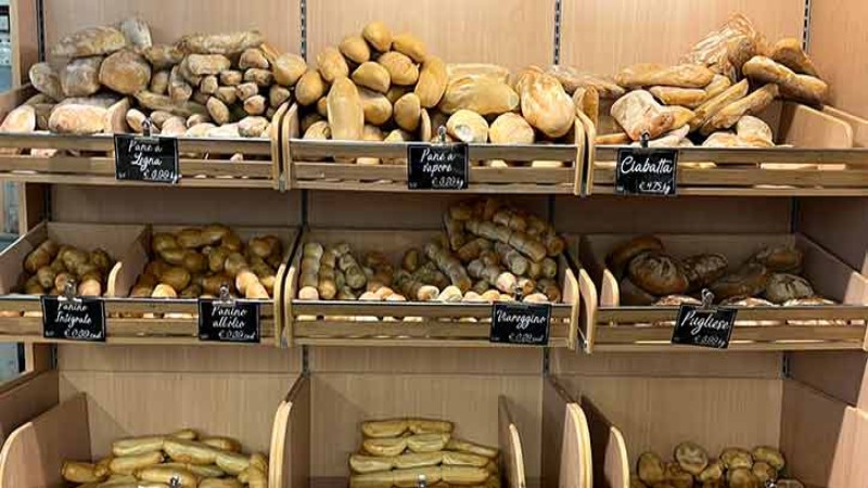 Espositore di panetteria con pane fresco, baguette e ciabatta su scaffali in legno.