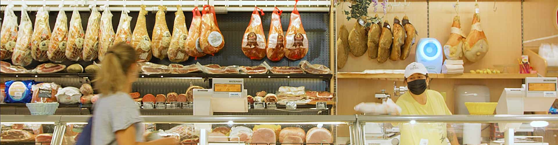 Banco gastronomia italiana con salumi, prosciutti appesi e personale al servizio dei clienti.