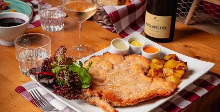Cotoletta di vitello impanata con patate arrosto, insalata e vino spumante su tavolo rustico.