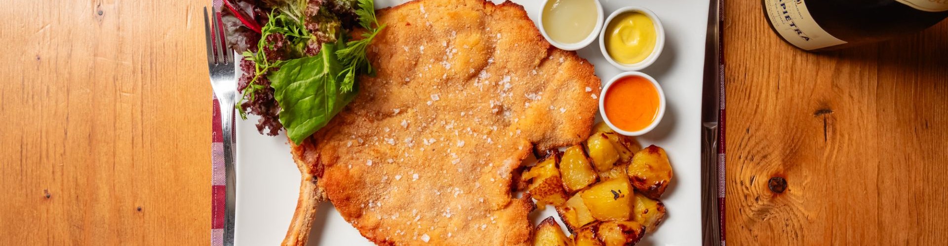 Cotoletta di vitello impanata con patate arrosto, insalata e salse su tavolo rustico in legno.