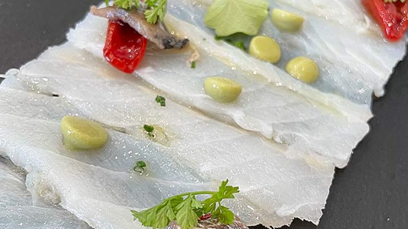 Sottili fette di carpaccio di baccalà con crema alle erbe, pomodorini e erbe fresche.