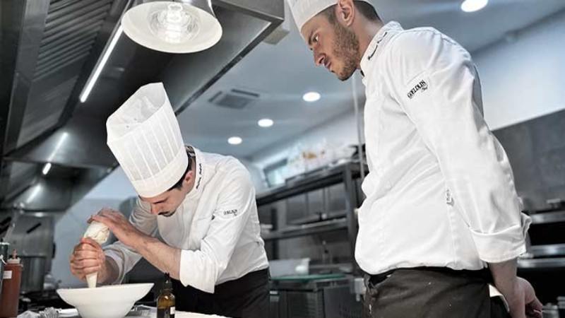 Chef che impiatta una portata mentre un collega osserva in una cucina professionale.