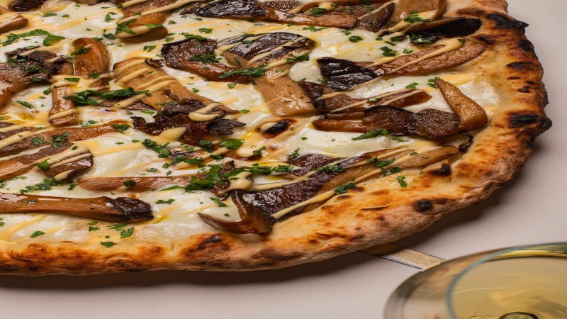 Pizza gourmet con funghi, formaggio fuso, erbe fresche e salsa accanto a un bicchiere di vino.