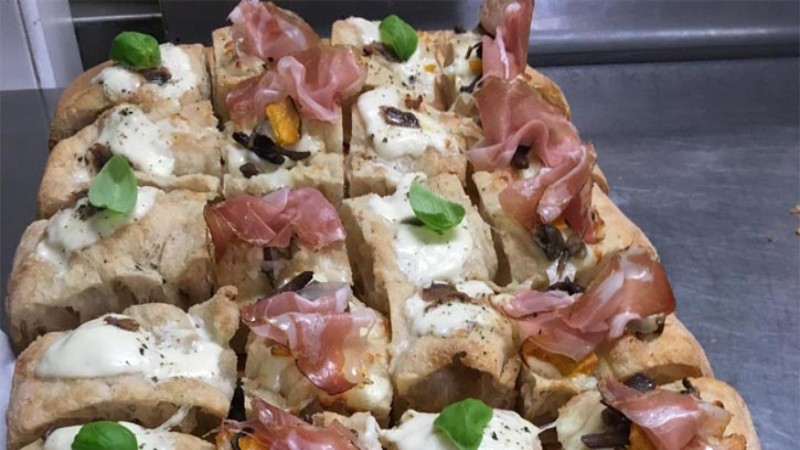 Tranci di focaccia con formaggio fuso, prosciutto crudo e foglie di basilico pronti da servire.