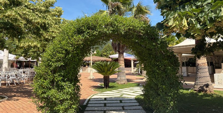 Terrazza con arco verde, palme e tavoli all’aperto sotto un cielo azzurro e sereno.