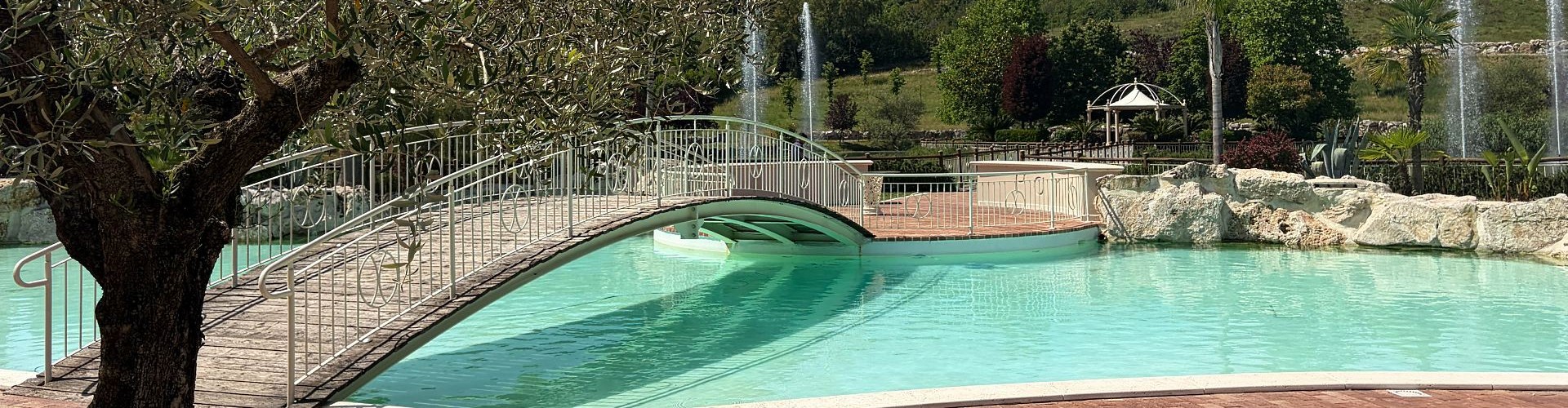 Piscina turchese con ponte ad arco, fontane e rigoglioso giardino mediterraneo sotto il cielo azzurro.