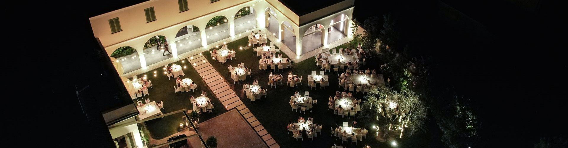 Vista aerea notturna di elegante evento gastronomico all’aperto in cortile illuminato.