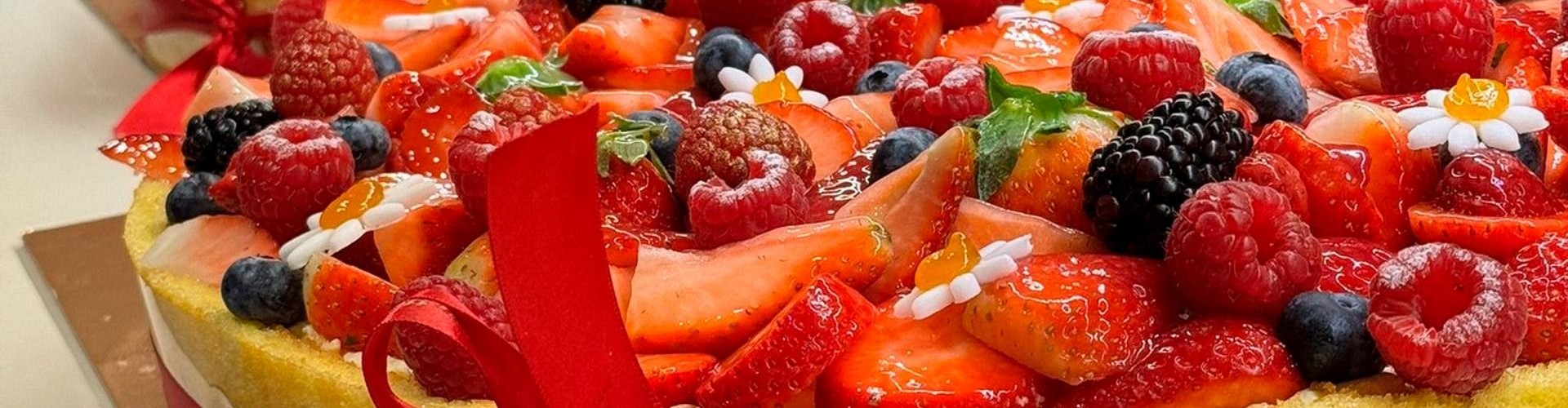 Torta a forma di cuore con frutti di bosco e nastro rosso, decorata con fragole e lamponi.