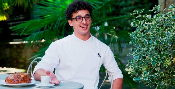 Pasticcere sorridente in divisa bianca seduto all'aperto con caffè e torta.