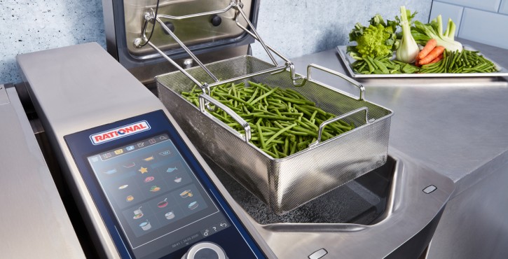 An overhead view of an iVario Pro 2S where one of the pans is being used to boil green beans in the autolift basket, which is about to lowered into the boiling water. A tray of other vegetables sits on the countertop in the background.