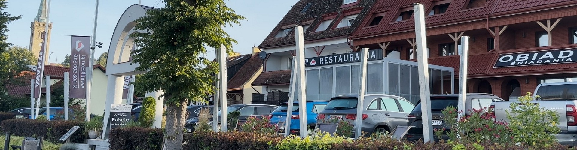 Widok na hotel i restaurację z zaparkowanymi samochodami, szyldami i wieżą kościoła w tle.