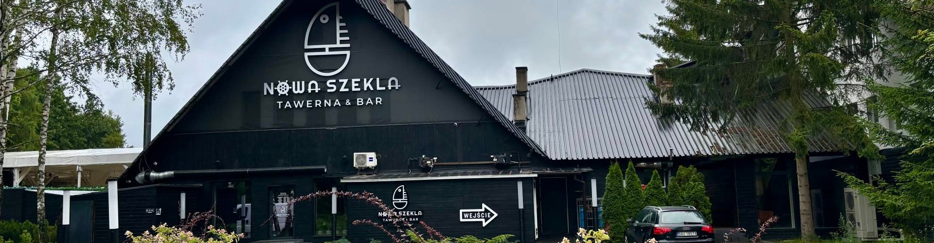 Widok frontowy Nowa Szekla Tawerna & Bar z nowoczesną czarną elewacją i logo ryby nad wejściem.