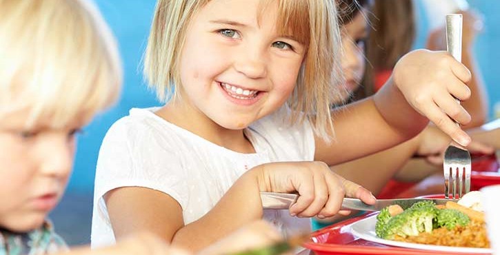 A young blond girl smiles at the camera while she uses her knife and fork to eat her school lunch on a red tray. There are other children around her also enjoying their lunch of rice, broccoli and chicken. 