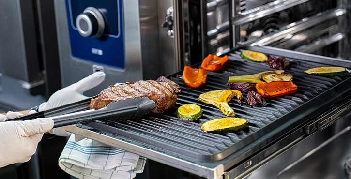Chef using tongs to remove grilled steak and vegetables from the iCombi Pro oven.