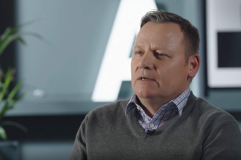 Mann im grauen Pullover spricht bei einem Interview in Innenraum mit unscharfem Hintergrund.