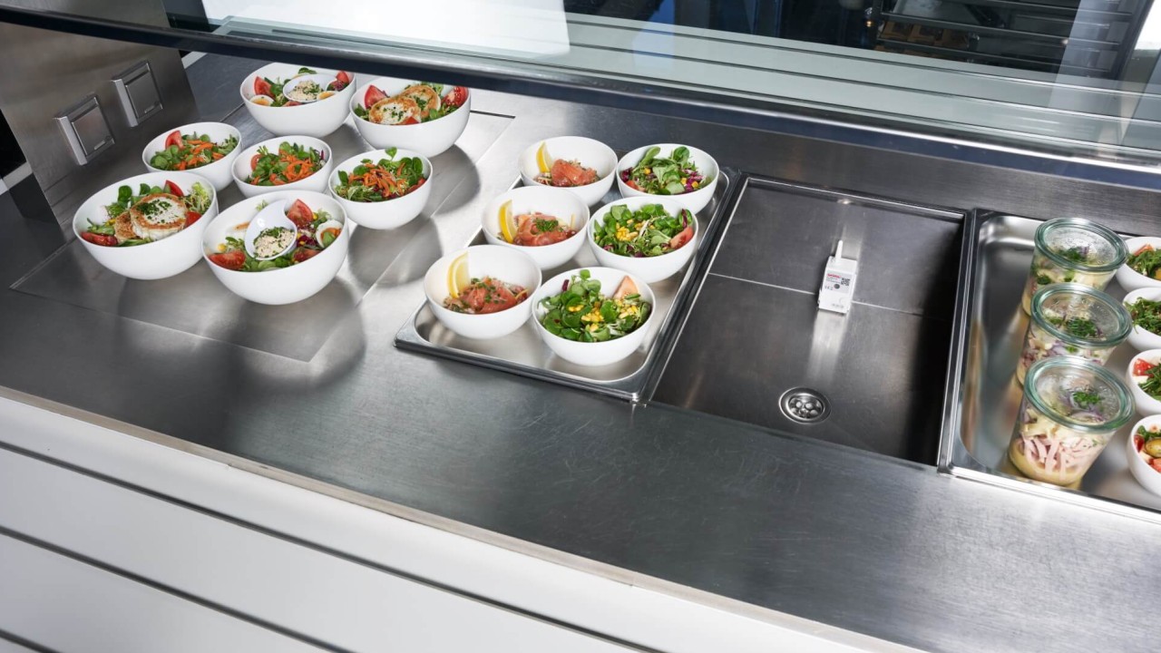 Fresh salads in bowls displayed on a stainless steel buffet counter with a small sensor.