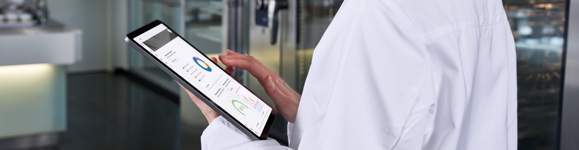 A woman in a white lab coat and hairnet looking at a tablet displaying data in a professional kitchen environment.