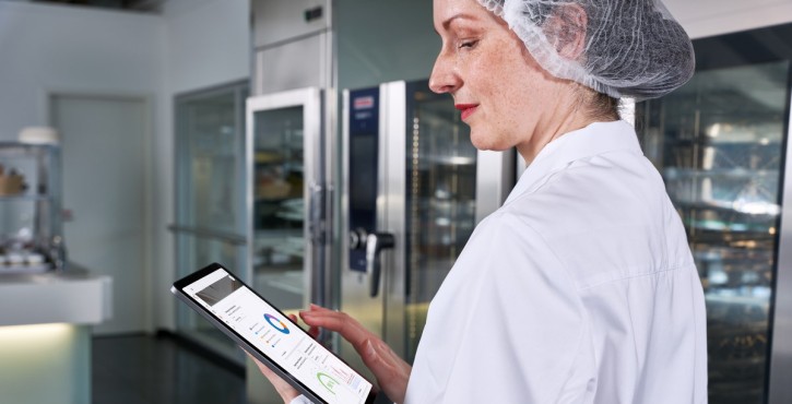 Eine Frau in einem weißen Laborkittel und Haarnetz, die ein Tablet mit Daten in einer professionellen Küchenumgebung betrachtet.