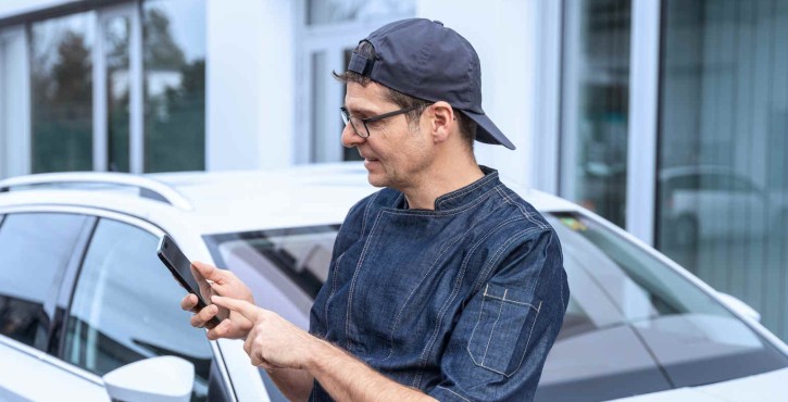 Un chef portant une veste de cuisine en denim et une casquette, debout à l'extérieur à côté d'une voiture blanche en consultant son téléphone.