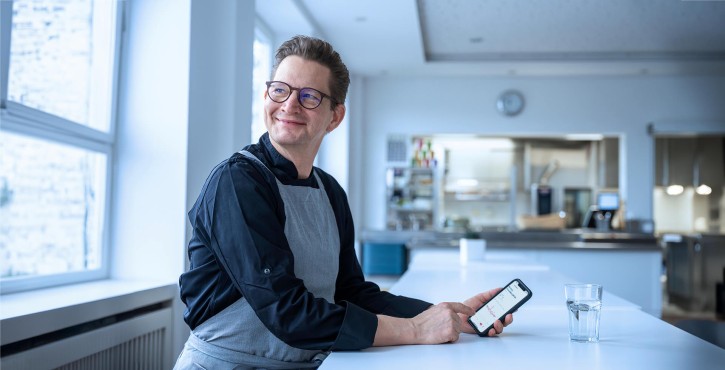 Ein zufriedener Koch sitzt an einem Tisch in der Küche, hält ein Smartphone mit der ConnectedCooking App.