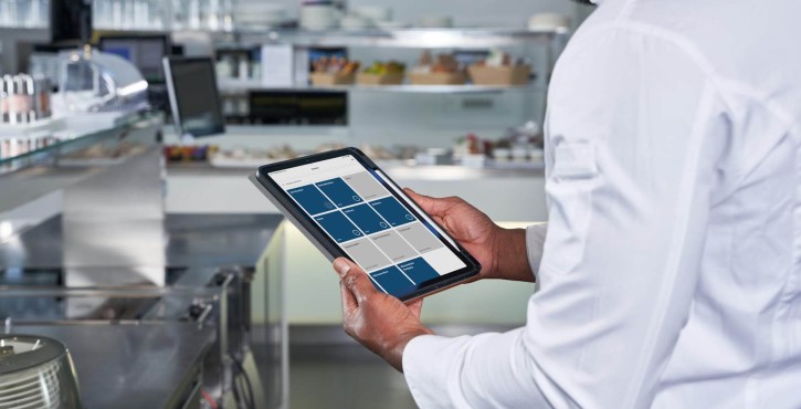 Ein Koch nutzt ein Tablet, um Aufgaben in einer Küche zu verwalten, mit einer modernen Küche im Hintergrund.