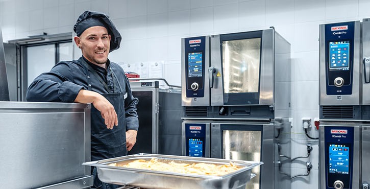 Un chef se tient à côté d'un four RATIONAL iCombi Pro, souriant, avec un plateau de nourriture devant lui et d'autres fours en arrière-plan.