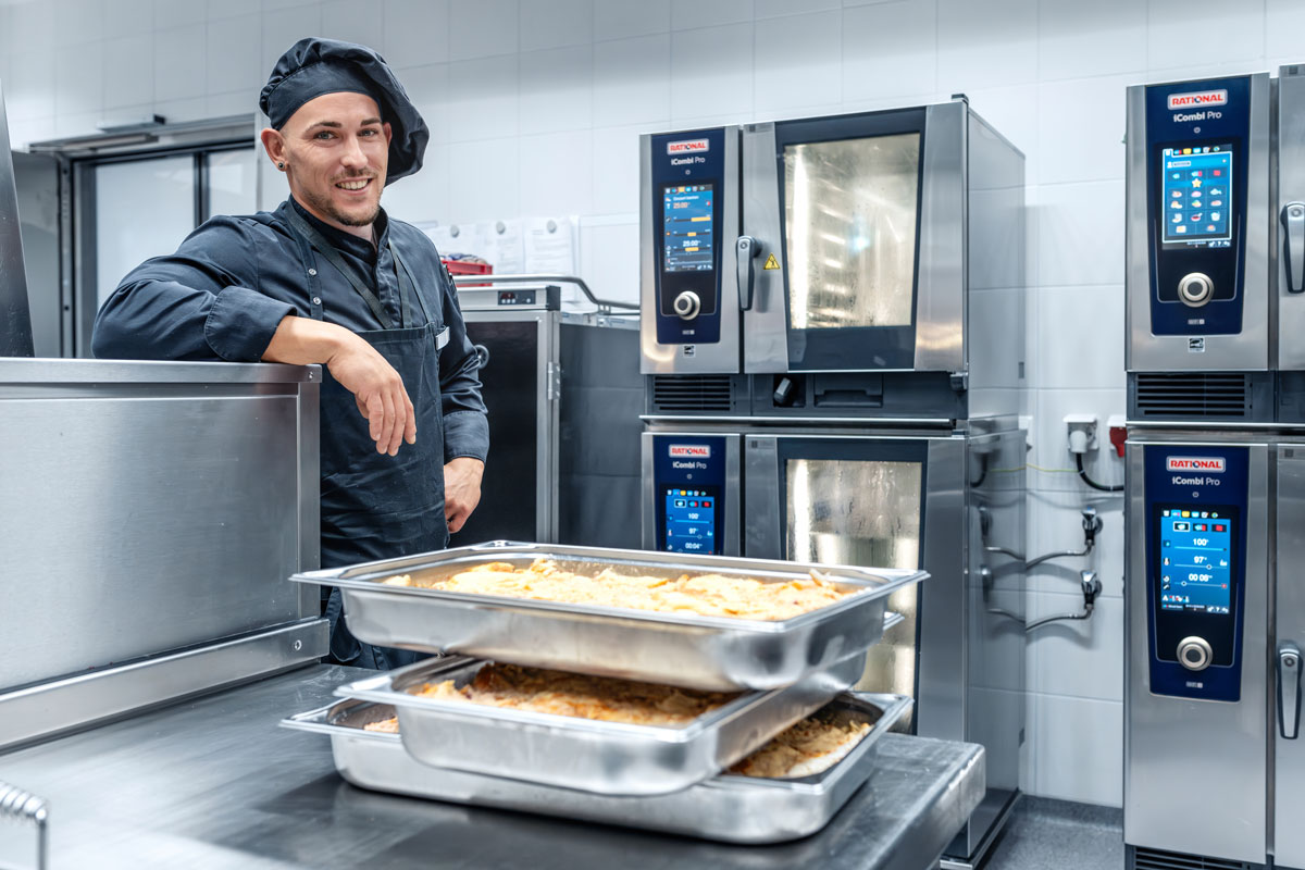 A chef stands next to a RATIONAL iCombi Pro oven, smiling, with a tray of food in front of him and other ovens in the background.