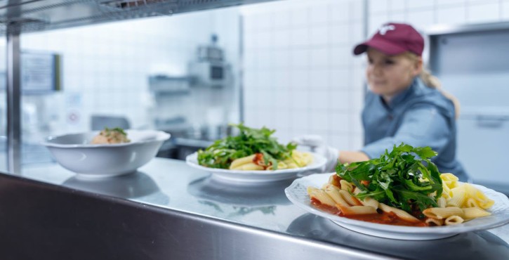 Eine Köchin stellt einen Teller Pasta mit Salat in einer Küche ab, mit zwei weiteren Tellern im Vordergrund.