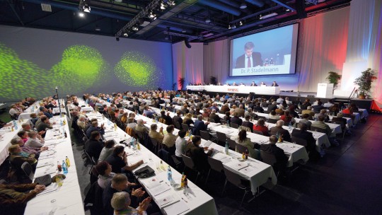 A large conference hall filled with attendees seated at tables, listening to a speaker presenting on a screen.