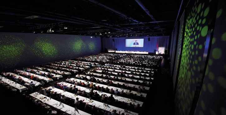 Large conference hall filled with attendees seated at long tables, watching a speaker on stage with a presentation screen above.