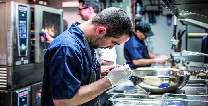 A chef in a blue uniform carefully prepares food in a professional kitchen, while other chefs work in the background.