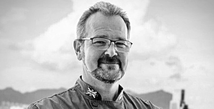 Black and white portrait of Andrew Twells in a chef’s jacket with glasses, in front of blurred buildings.