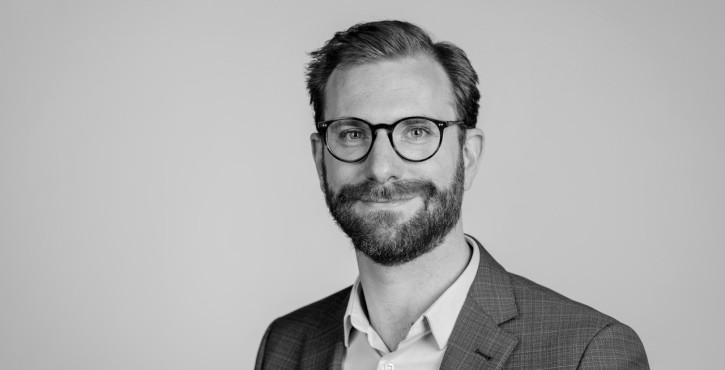 A man with glasses and a beard smiling in a portrait wearing a blazer and shirt, in black and white.