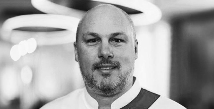 A man with a short beard smiling in a portrait, wearing a chef's uniform, in black and white.